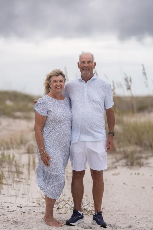 Two adults in white and patterned blue stand side by side on the beach at Vilano Beach, smiling at one another with dunes in the background.