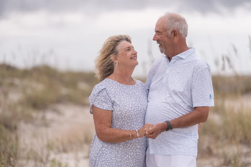 A couple in white and patterned blue face each other while holding hands on the sand at Vilano Beach, with beach vegetation behind them.