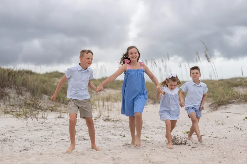 Four children hold hands while walking across the sand at Vilano Beach, wearing shades of blue and white, with dunes visible in the distance.