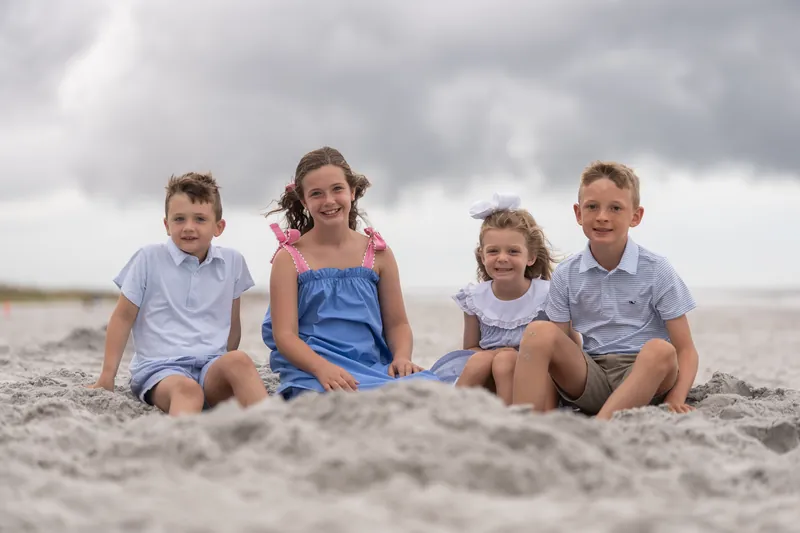 Four children sit nestled together in the sand, two in light blue shirts and two in darker blue outfits.