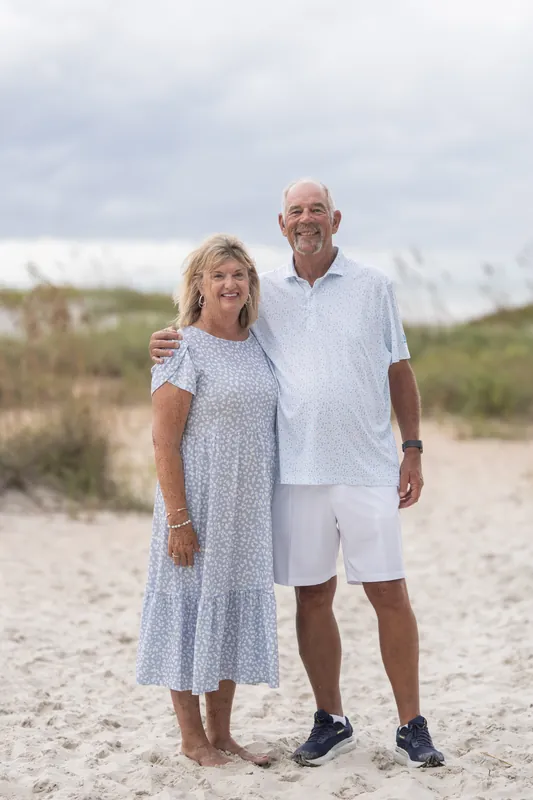 Kimberly Davis and partner stand arm-in-arm on the sand at Vilano Beach, dressed in light layers with dune grass behind them.