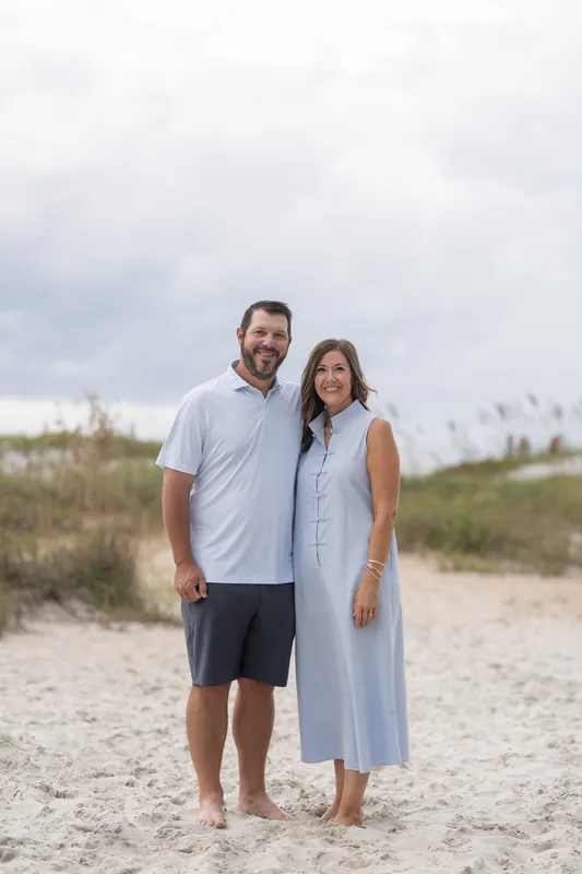 Two adults stand close together on the beach at Vilano Beach with dunes and beach grass visible in the background.