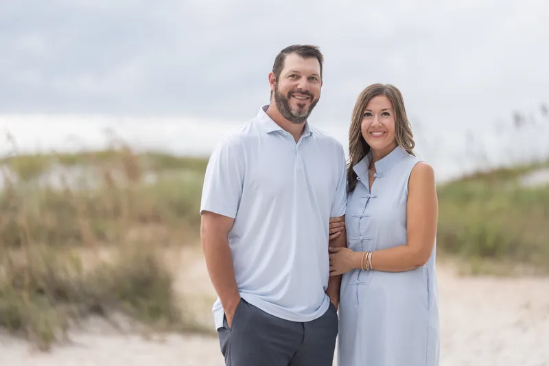 A couple smiles at the camera on the sand at Vilano Beach, with coastal dunes and vegetation framing the scene behind them.