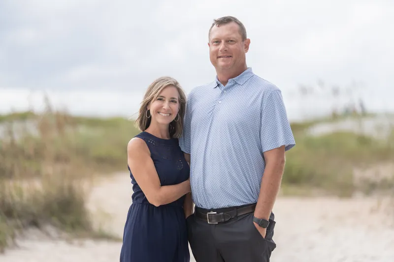 Two people stand together on the sand at Vilano Beach with dune grasses and overcast sky behind them.