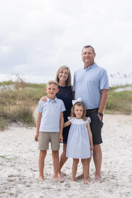 A family of four stands barefoot on Vilano Beach with beach grass and overcast sky in the background.