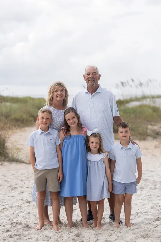 Five family members of varying heights stand barefoot together on the beach with sea oats and cloudy sky in the distance.