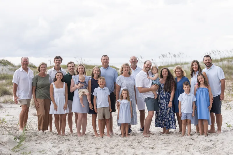 Extended family of fifteen gathers on the sand at Vilano Beach with dune grasses behind them under an overcast sky.