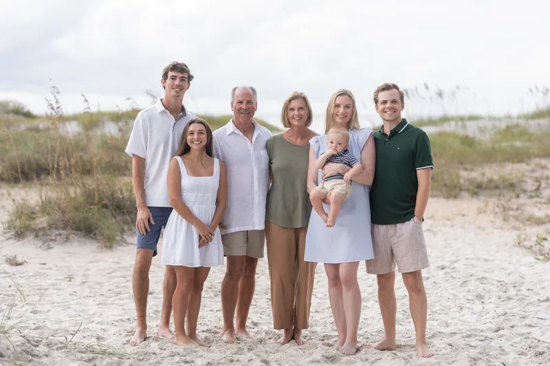 Six family members pose barefoot on the sand at Vilano Beach, one holding a small child, with marsh grasses in the background.