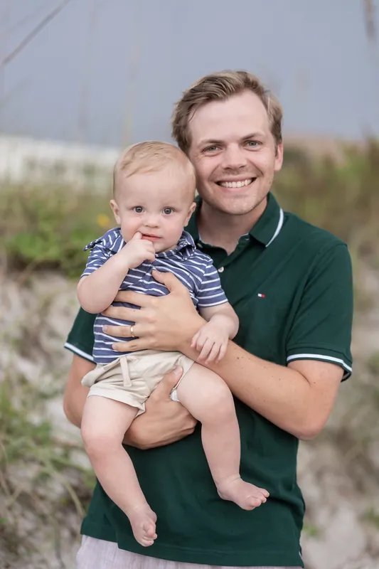 A man in a green polo shirt holds and smiles at a baby wearing a striped shirt, with beach dunes behind them.