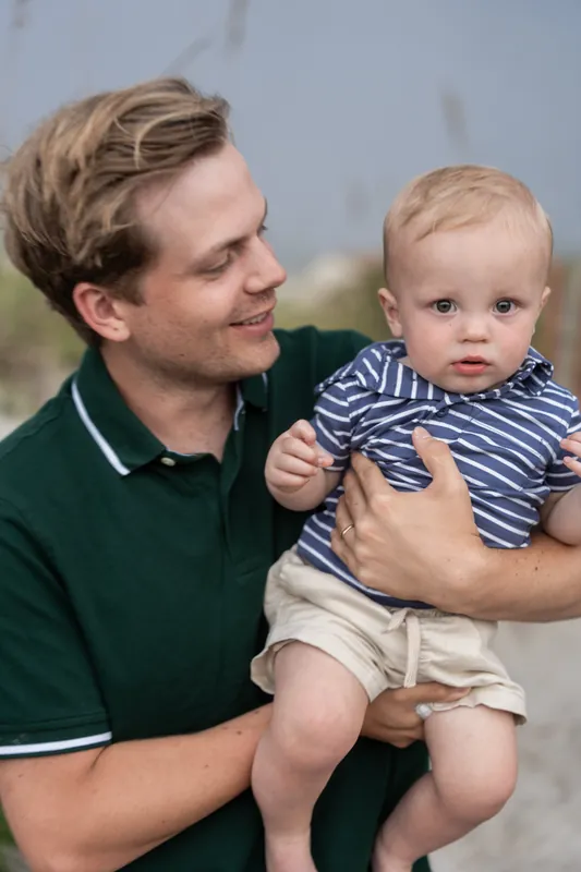 A man in profile gazes at the toddler he's holding, both wearing striped and solid colored shirts near the shoreline.