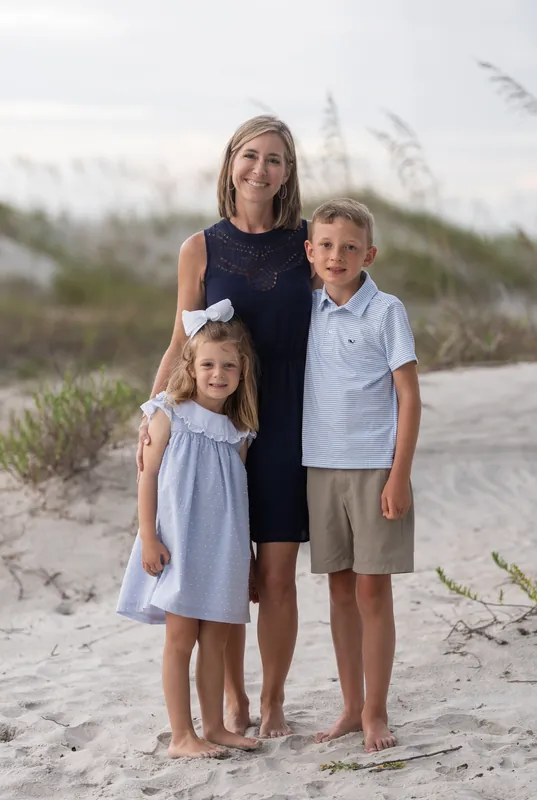 A woman in a black sleeveless dress stands with two children in light blue and khaki on Vilano Beach's sandy path lined with sea oats.