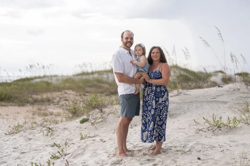 A man in a white shirt holds a toddler while standing beside a woman in a navy floral dress on the beach.