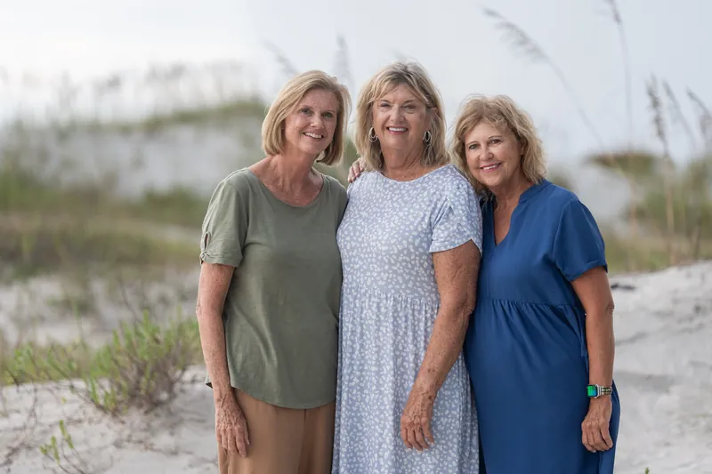 Three women in olive, lavender, and navy stand together on the sand with dune vegetation behind them at Vilano Beach.