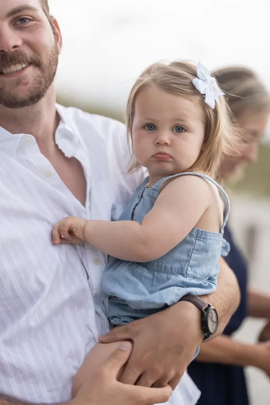 A man in a white shirt holds a small child with a white bow while smiling at the camera on the beach.