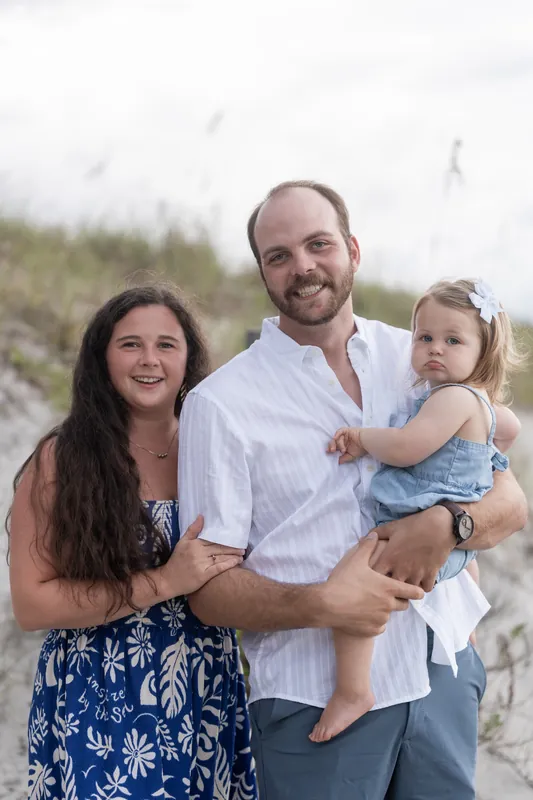 Two adults and a young child in a light blue outfit pose together on the sandy shore with sea grass visible behind them.