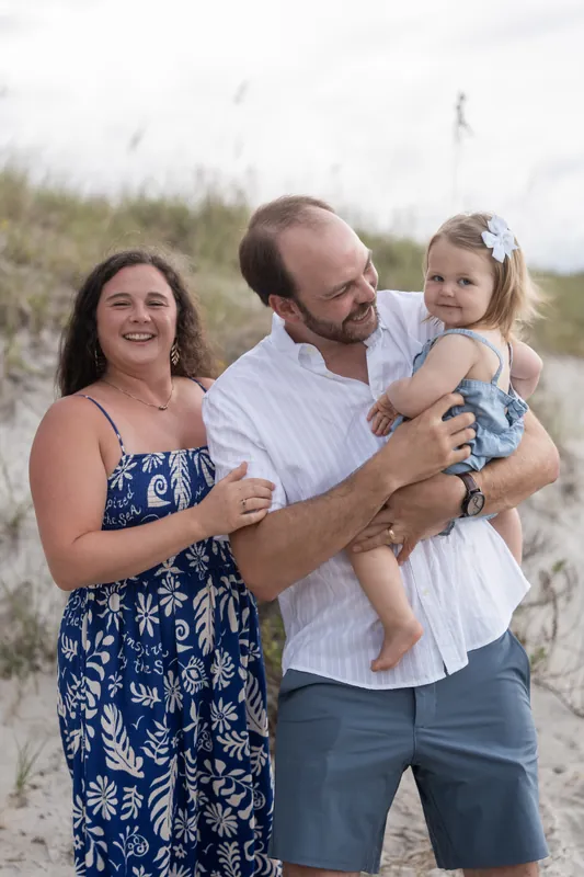 A couple stands close together holding their small child in a white bow on Vilano Beach's sandy expanse.
