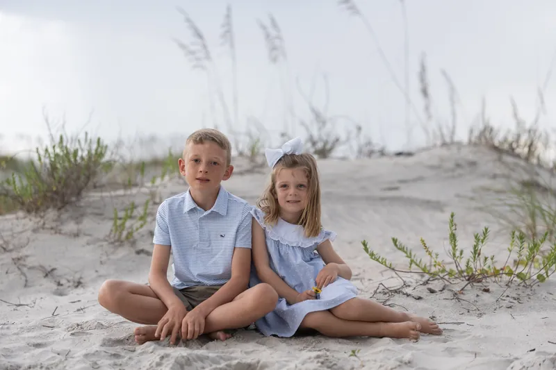 Two children in matching light blue shirts sit barefoot on sand at Vilano Beach with sea oats behind them.