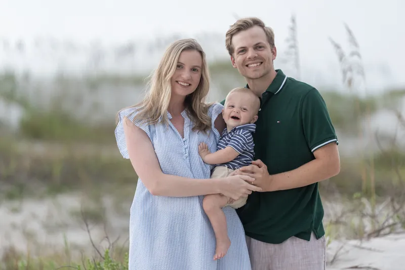 Two adults hold a small child on sand dunes at Vilano Beach with sparse beach grasses visible.