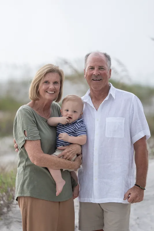 Two older adults hold a toddler on the beach at Vilano Beach, dunes and sea oats in the background.