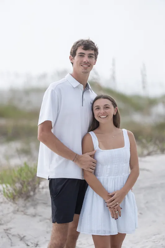 A young man in a white polo and a young woman in a white dress stand together on sand at Vilano Beach.