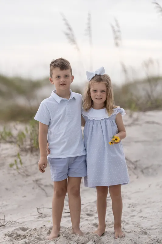 A boy in a light blue shirt and a girl in a pale blue dress stand side-by-side on the beach, both barefoot.
