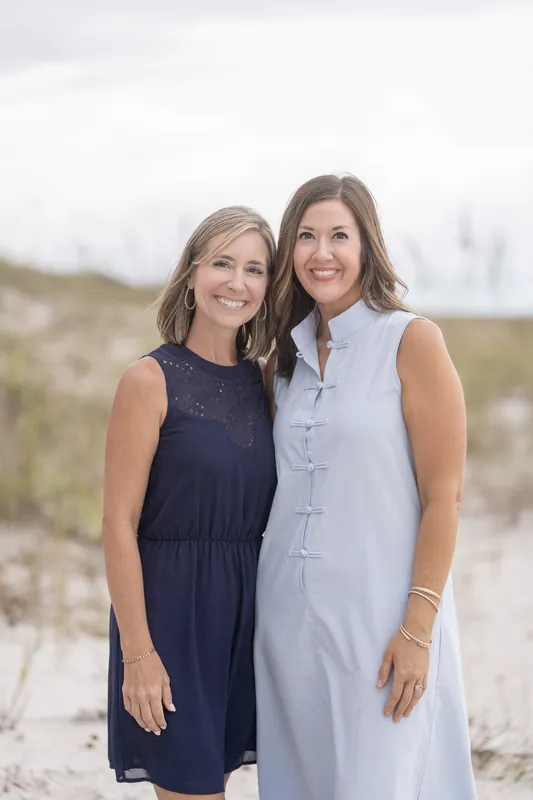 Two women pose together on the beach with dune grasses behind them, one wearing a navy sleeveless dress and the other in a light blue linen shift.