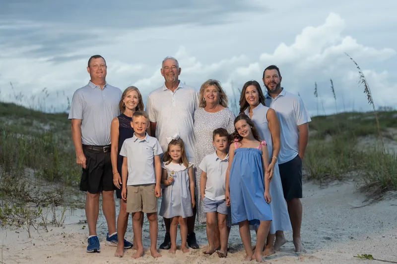A multi-generational family group stands together on sand with beach grasses and dunes visible behind them.