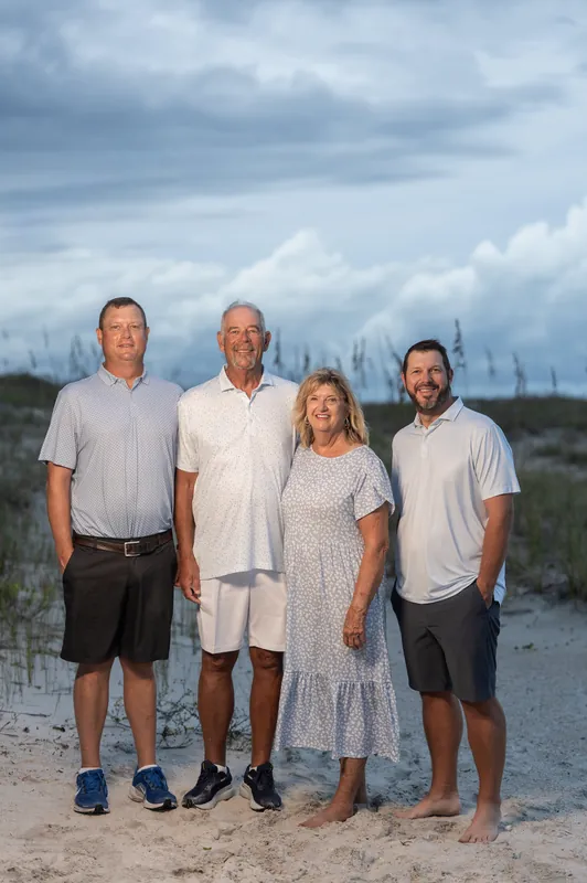 Four adults stand together on the beach wearing neutral tones, with sea oats and overcast sky visible in the background at Vilano Beach.