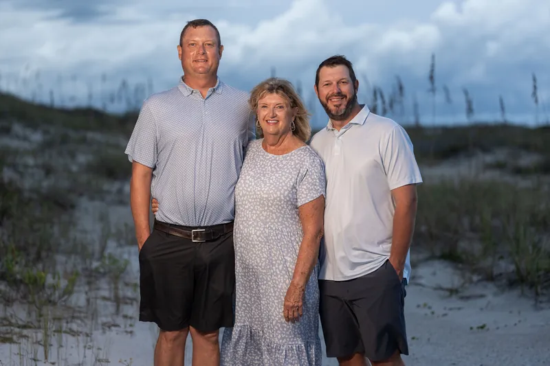 Three adults stand on the sand at Vilano Beach—two men flanking a woman in the center, all wearing light-colored casual shirts.