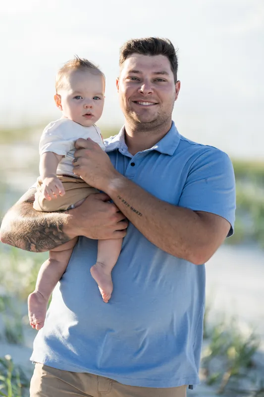 Man in blue polo holds smiling toddler at St. Augustine Beach with dunes and beach grass behind them.
