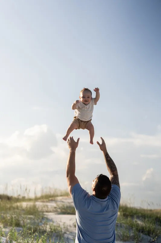 Father tosses laughing toddler in the air on the sand at St. Augustine Beach on a clear day.