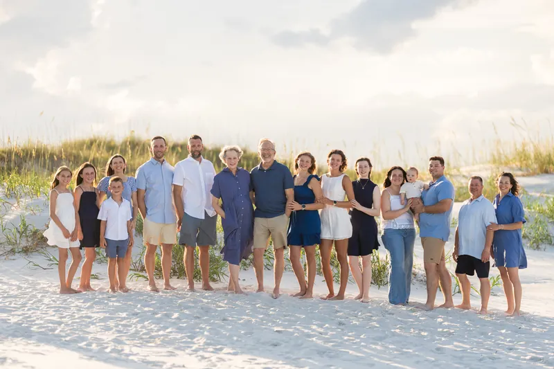 Large family group of fifteen poses barefoot on the beach at St. Augustine Beach with dune grasses visible.