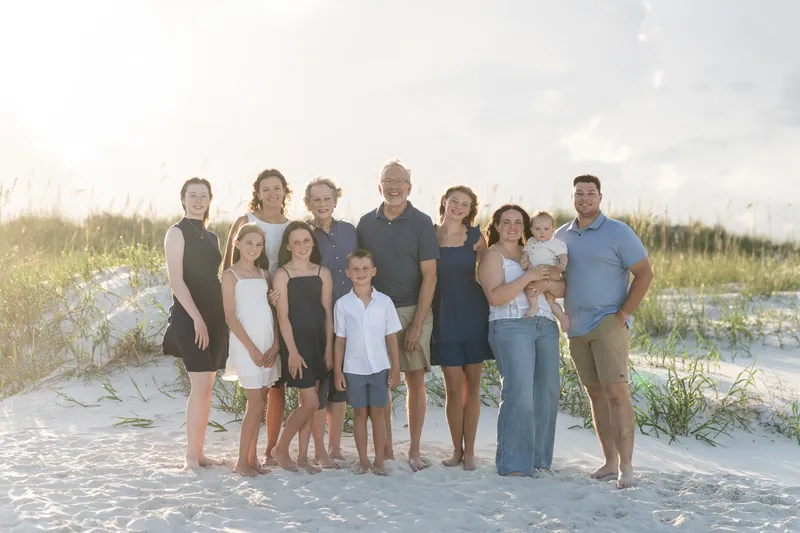 Multi-generational family of thirteen stands close together on the sand with coastal dunes in the background.