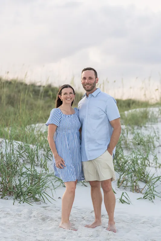 A man and woman stand barefoot on the sand at St. Augustine Beach, with dune grass behind them under an overcast sky.