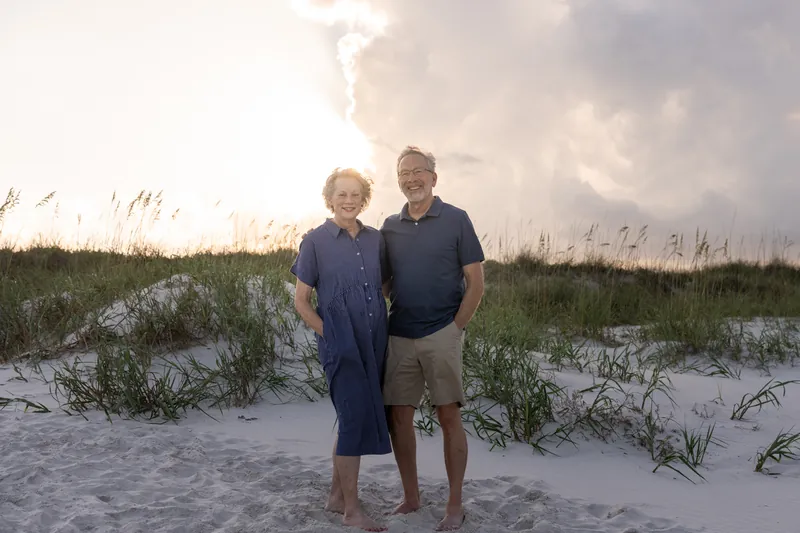 Two family members pose together on the beach with sand dunes and sea grass visible in soft evening light.