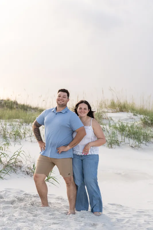 A man in a blue shirt and woman in jeans stand with hands on hips on the beach, dunes behind them.