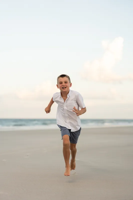 A young boy in a white shirt runs barefoot across the wet sand at St. Augustine Beach, smiling as waves roll behind him.