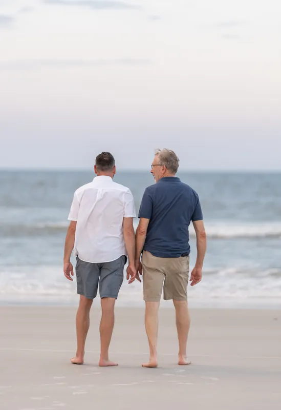 Two men walk barefoot away from the camera toward the ocean at St. Augustine Beach, their backs to the lens as gentle waves