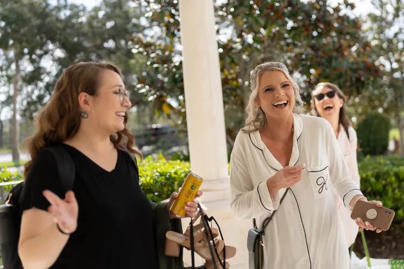 Kimberly laughs while holding a champagne bottle on a covered patio at River House Events, surrounded by friends in casual