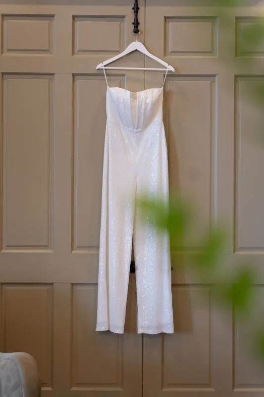 A white sleeveless jumpsuit hangs on a wooden hanger against paneled doors at River House Events.