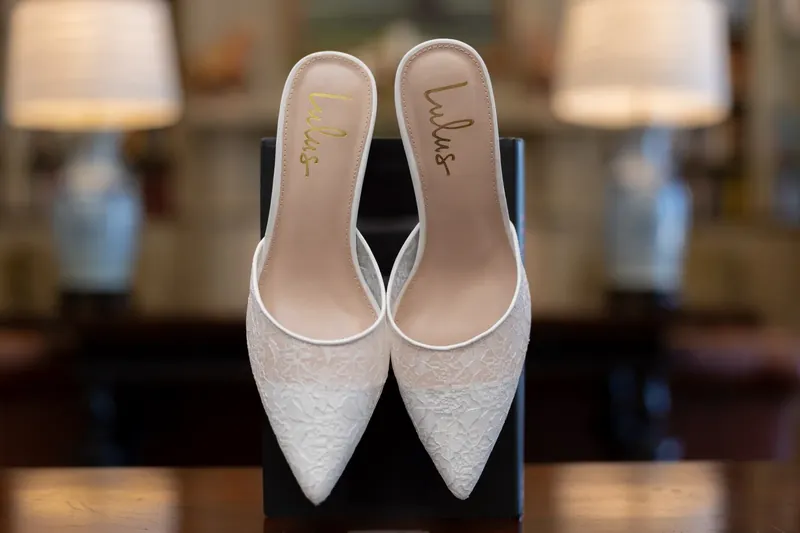 Pair of white pointed-toe heels with "Kimberly" written on the soles, displayed on a dark surface with lamps blurred behind.