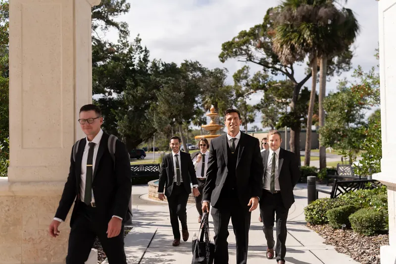 Landon and five groomsmen in black suits walk along a covered outdoor corridor with palm trees and manicured landscaping