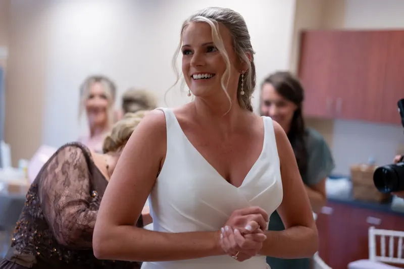 Kimberly smiles brightly in a white sleeveless dress while standing indoors at River House Events with guests blurred