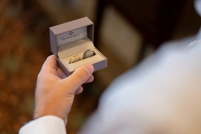 A hand holds open a small ring box displaying two gold wedding bands inside.