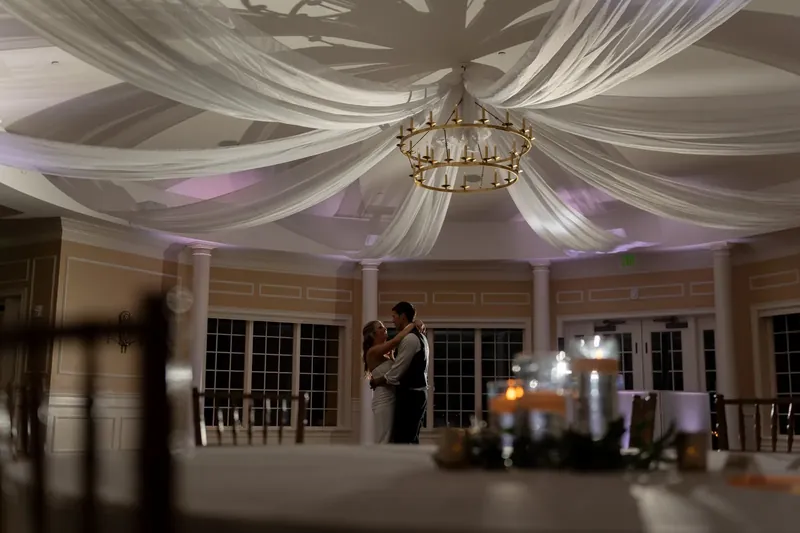 A chandelier hangs from draped fabric overhead in an elegant reception hall with tall windows.