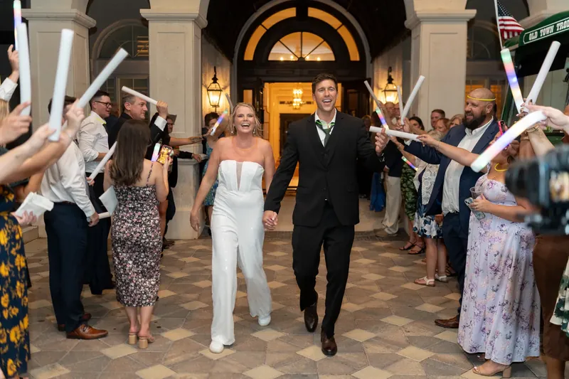 Kimberly in a white sleeveless gown and Landon in a dark suit walk through a ribbon tunnel as guests celebrate on a columned