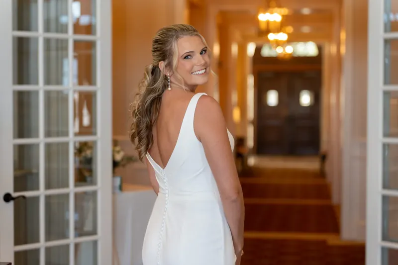 Kimberly turns back over her shoulder in her white V-neck wedding dress, standing in a warm-lit hallway at River House