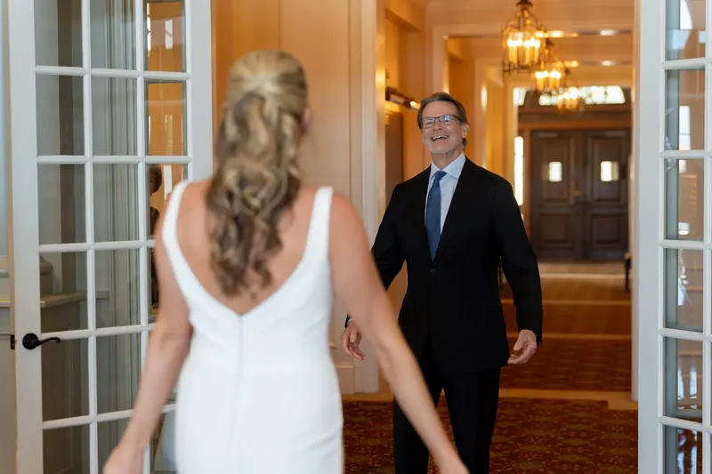 Landon walks toward Kimberly down a brightly lit corridor at River House Events as she faces away in her white wedding dress.