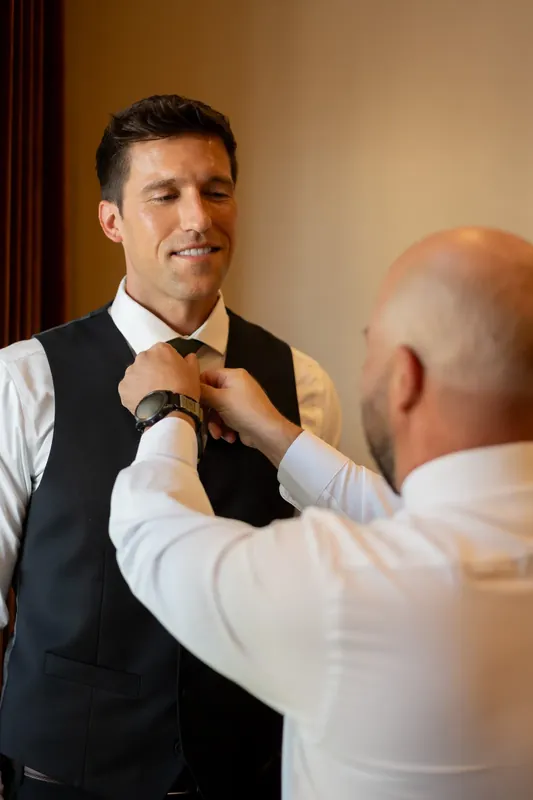 Landon smiles while someone adjusts his tie in a cream-colored room at River House Events.