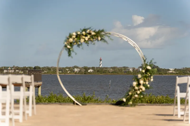 A circular white arbor decorated with white and green florals frames the water at River House Events.